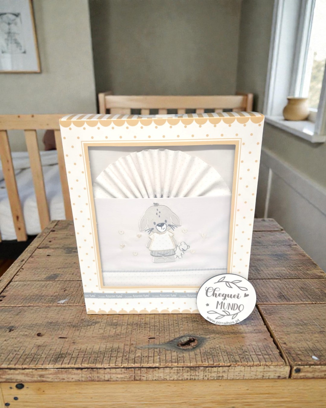 Caixa de manta ou roupa de bebé branca com bordado de cão e caixa com pontos bege sobre mesa de madeira.