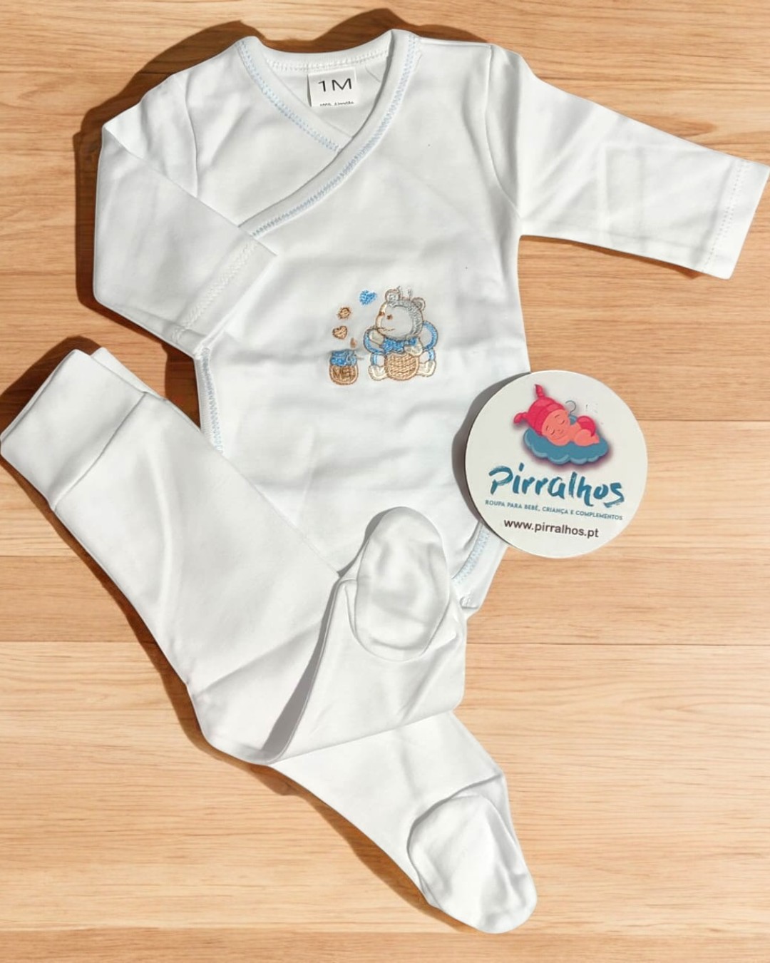 Conjunto de roupa de bebé branco com estampa de hipopótamo e ursinhos, etiquetas visíveis, sobre piso de madeira clara