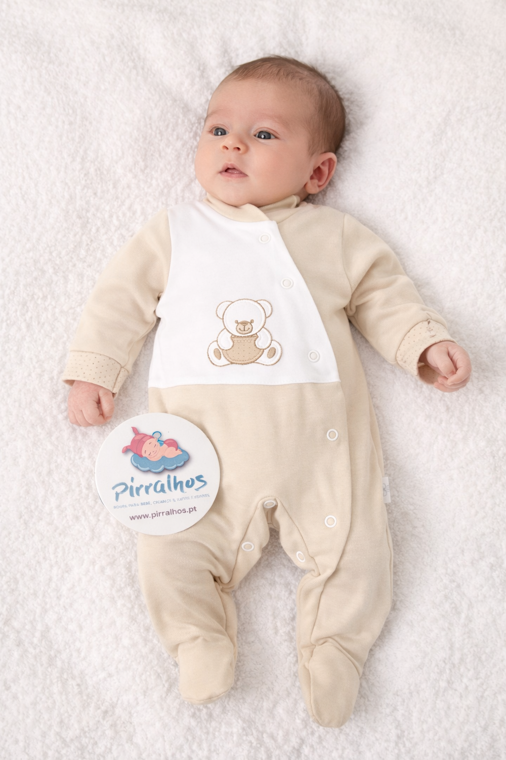 Bebé com macacão bege e branco com ursinho bordado e botões de pressão, sobre fundo branco macio