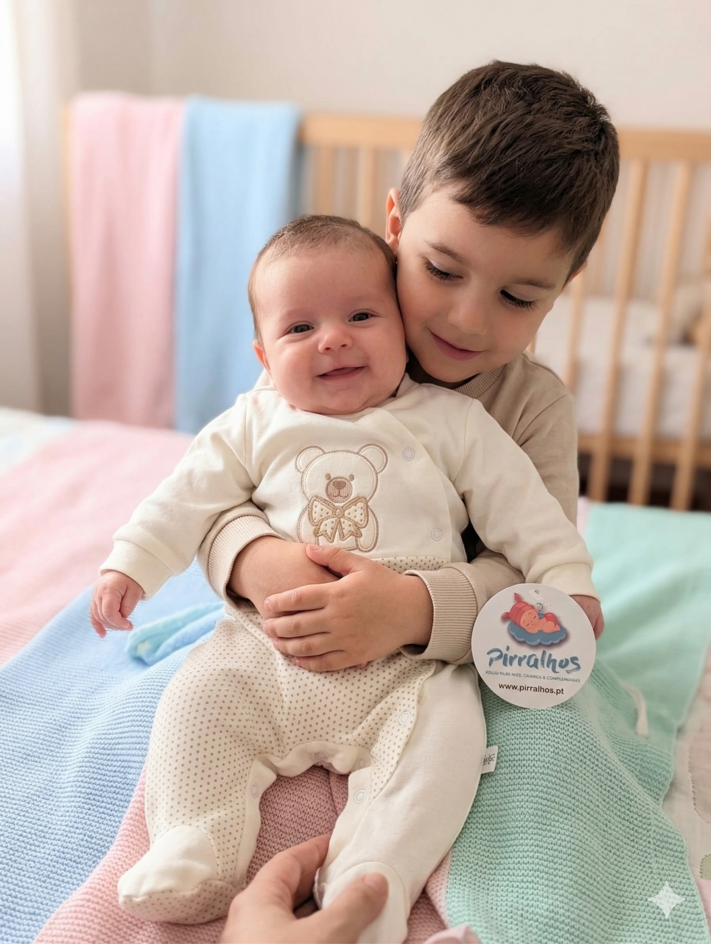 Bebé em macacão creme com ursinho bordado no colo de criança maior, mantas coloridas e berço ao fundo