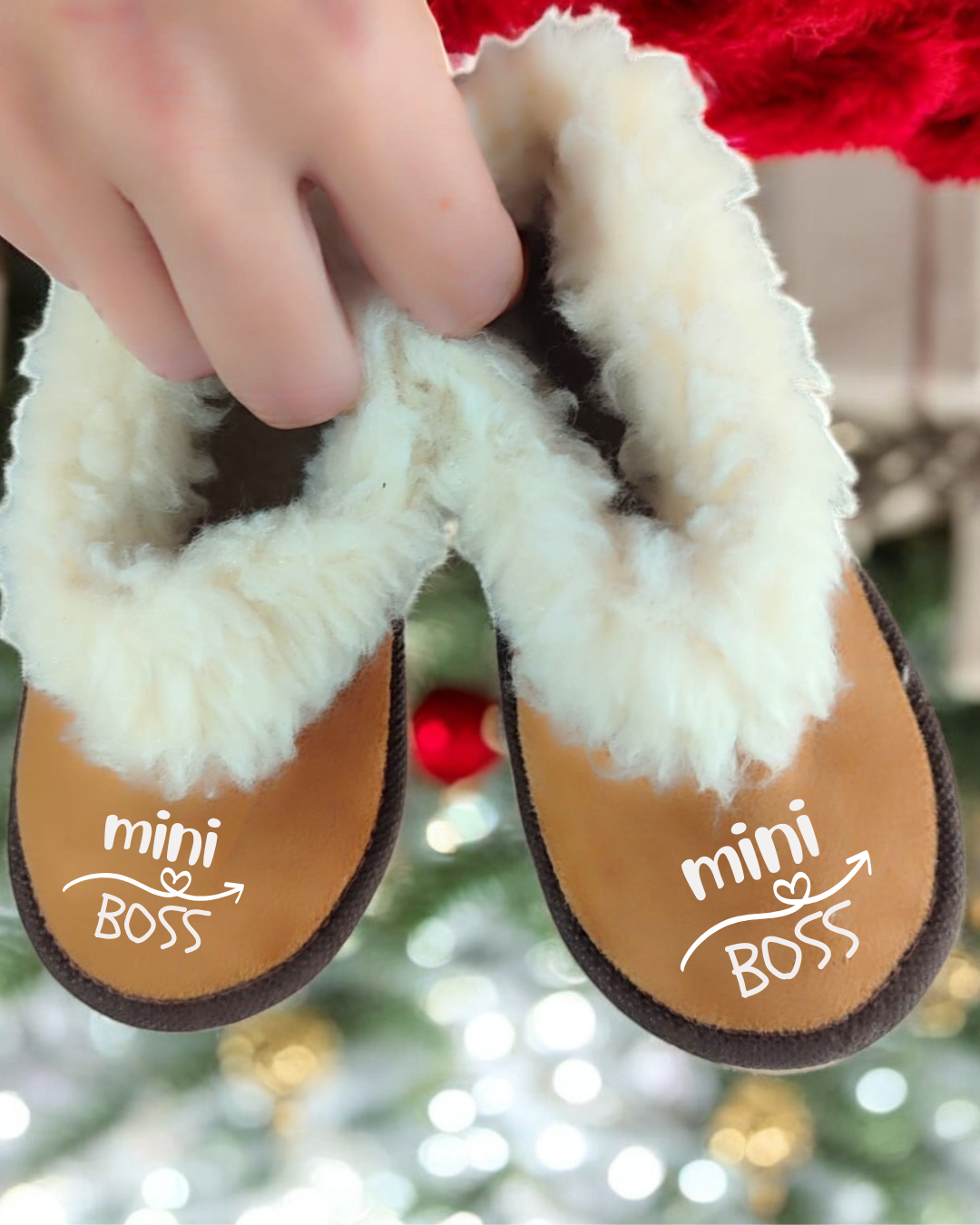 Pantufas marrons com pelúcia branca e texto mini BOSS, seguradas por uma mão com luzes de Natal ao fundo