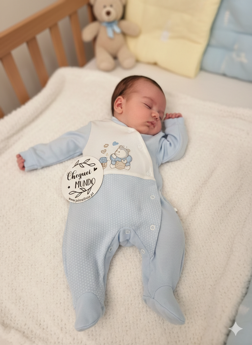 Bebé vestido com macacão azul e branco com desenho de ursinho a dormir numa cama com peluche castanho.
