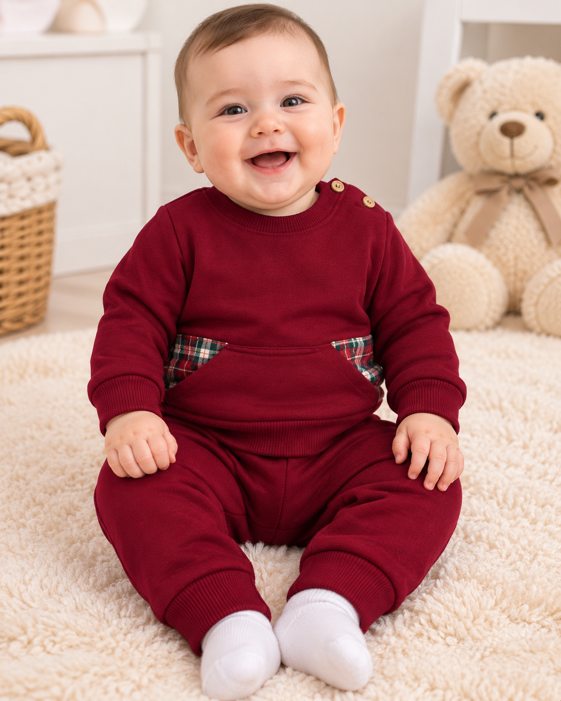 Bebé com fato vermelho bordô e detalhes xadrez sentado em tapete peludo claro com urso de peluche ao fundo.