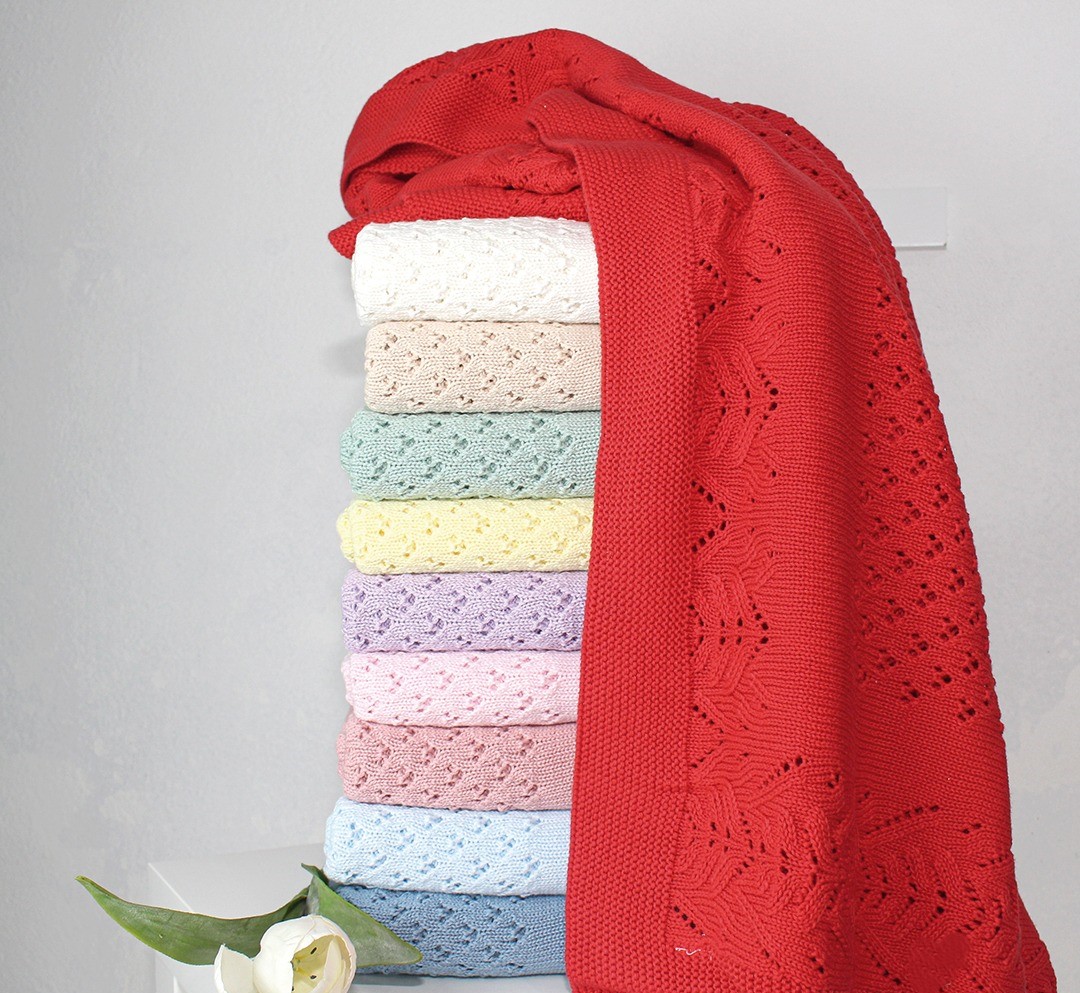 Empilhamento de mantas coloridas de tricot com padrões perforados e flor branca ao lado.
