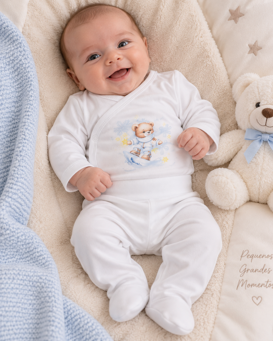 Bebé sorridente vestido com macacão branco com ursinho estampado, deitado em manta bege com cobertor azul e ursinho de peluche ao lado.