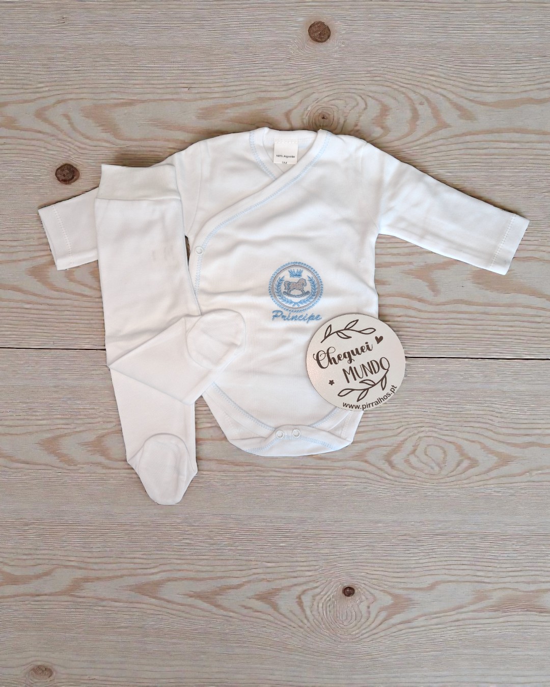 Conjunto de roupa de bebé branco com bordado azul sobre superfície de madeira clara