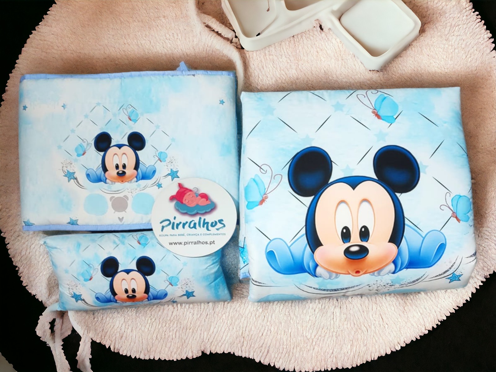 Três artigos de tecido azul claro com estampas do personagem Mickey Mouse colocados sobre uma superfície macia cor de rosa