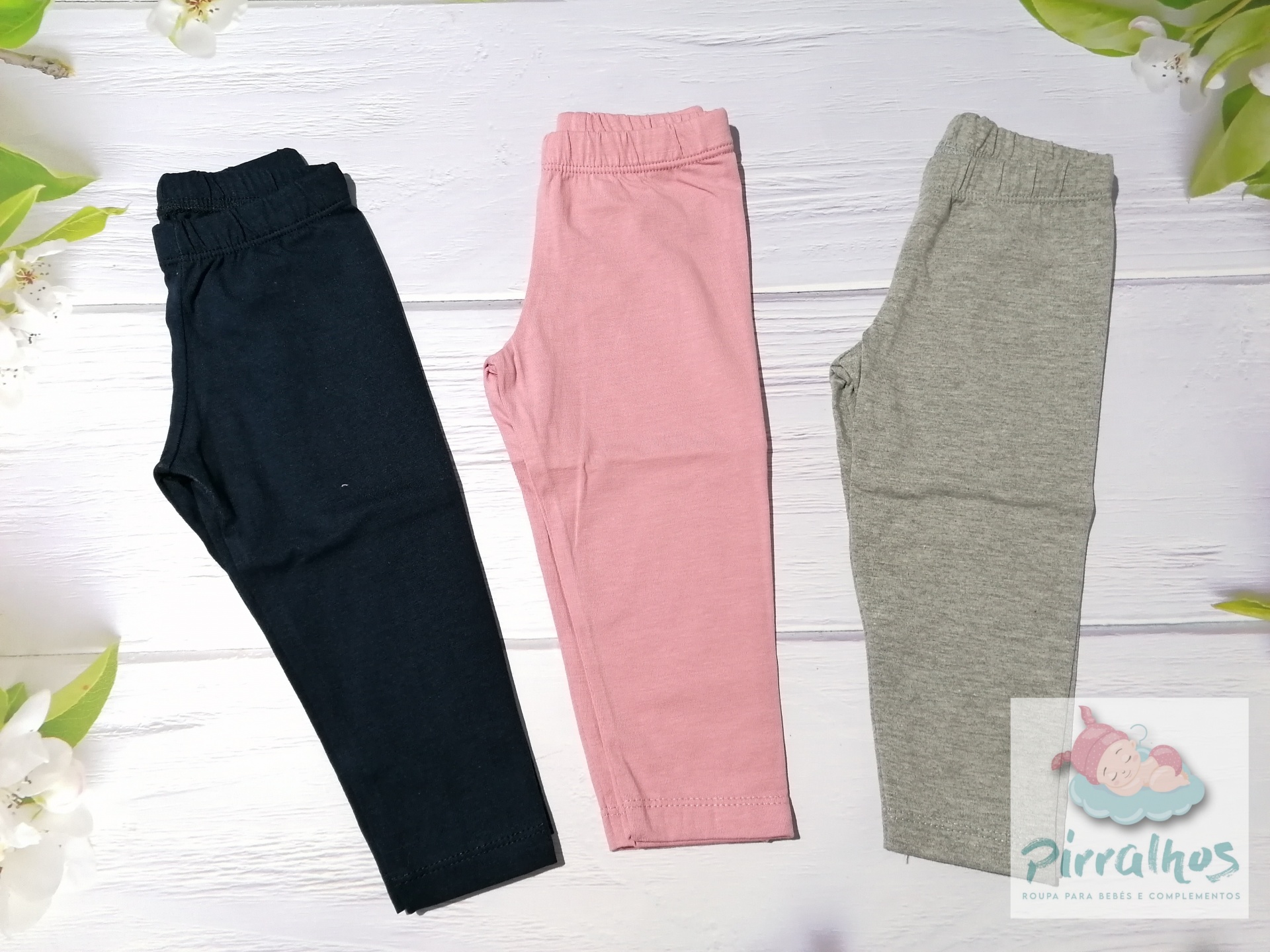 Três calças de criança em preto, rosa e cinza, em superfície branca de madeira com decoração floral