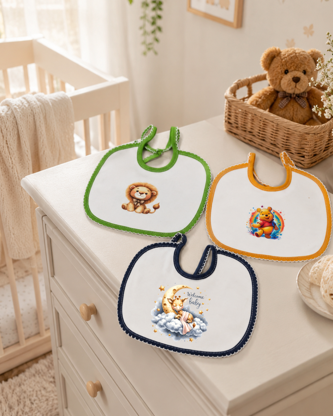 Três babetes brancos com desenhos infantis e bordas coloridas sobre uma cómoda branca com um urso de peluche na cesta ao lado.