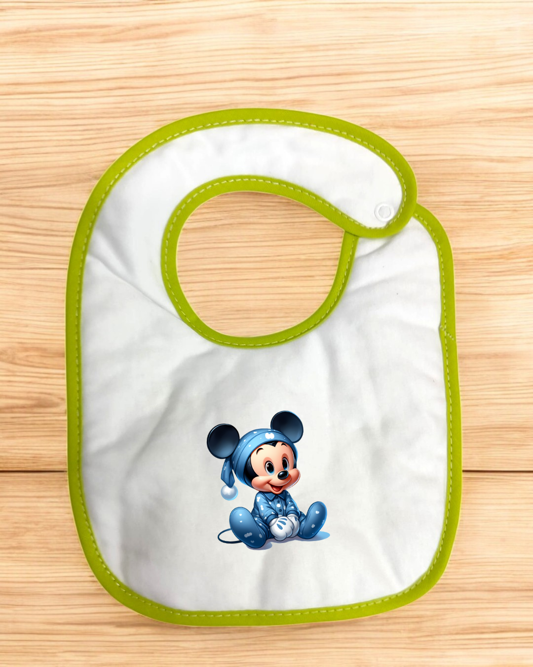 Babete branco com bordas verdes e desenho do rato Mickey em pijama azul sobre superfície de madeira clara