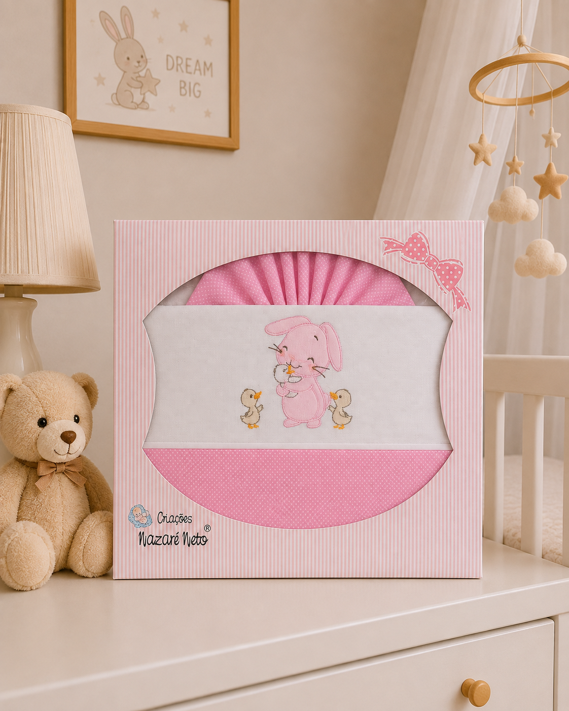 Roupa de cama rosa para bebé embalada em caixa decorada com coelhinho e patos, ao lado urso de peluche castanho.