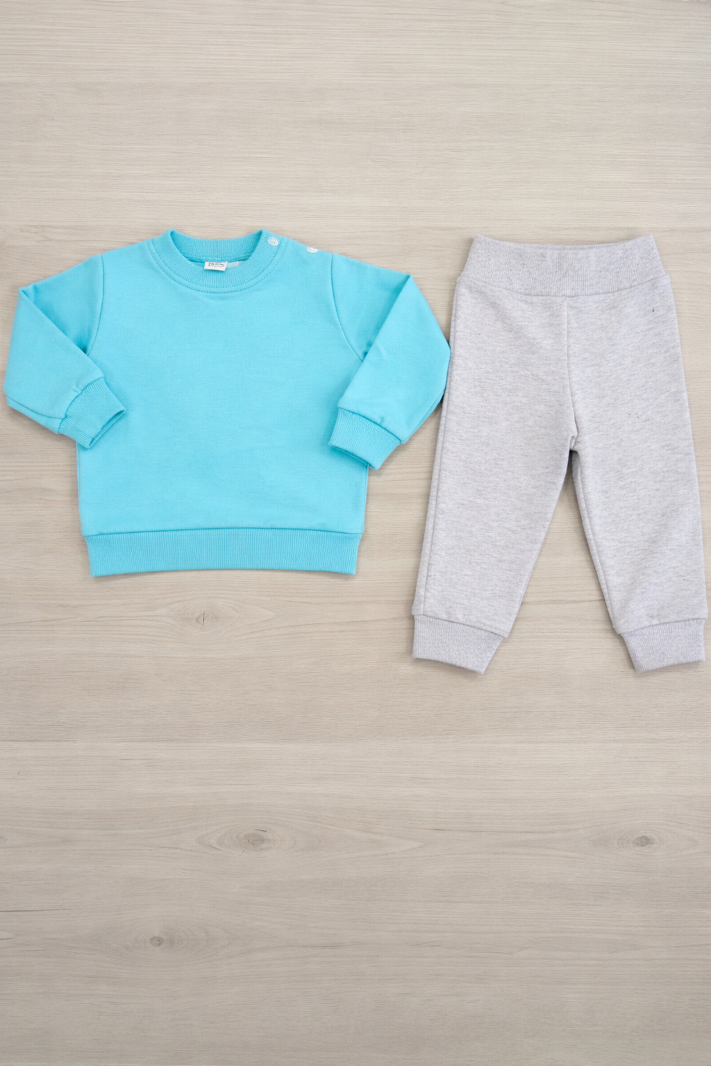 Conjunto de roupas infantis azul e cinza claro sobre chão de madeira claro.