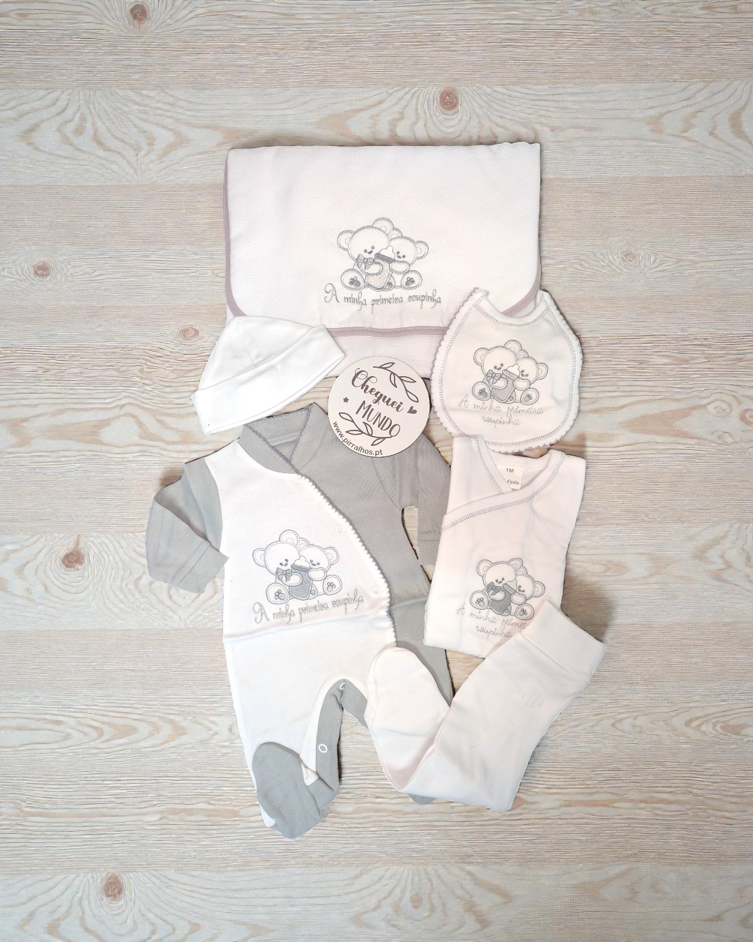 Conjunto de roupa para bebé branco e cinza com bordado de ursinhos e texto em tecido, exposto sobre fundo de madeira clara.