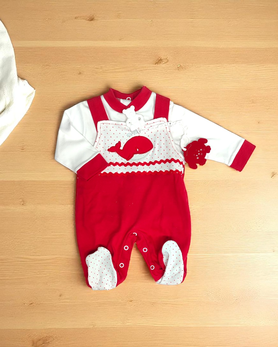 Macacão de bebé vermelho e branco com padrão de bolinhas e baleia, sobre fundo de madeira