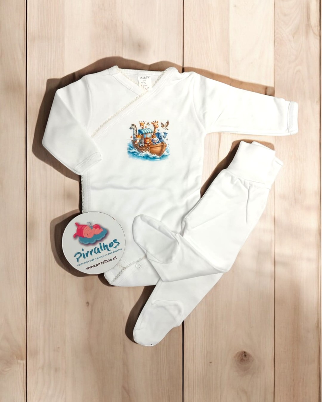 Conjunto roupa de bebé branco com ilustração de barco e animais sobre chão de madeira.