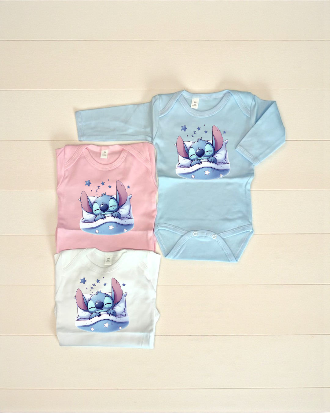 Três bodys de bebé azul, rosa e branco com estampa de personagem azul a dormir.