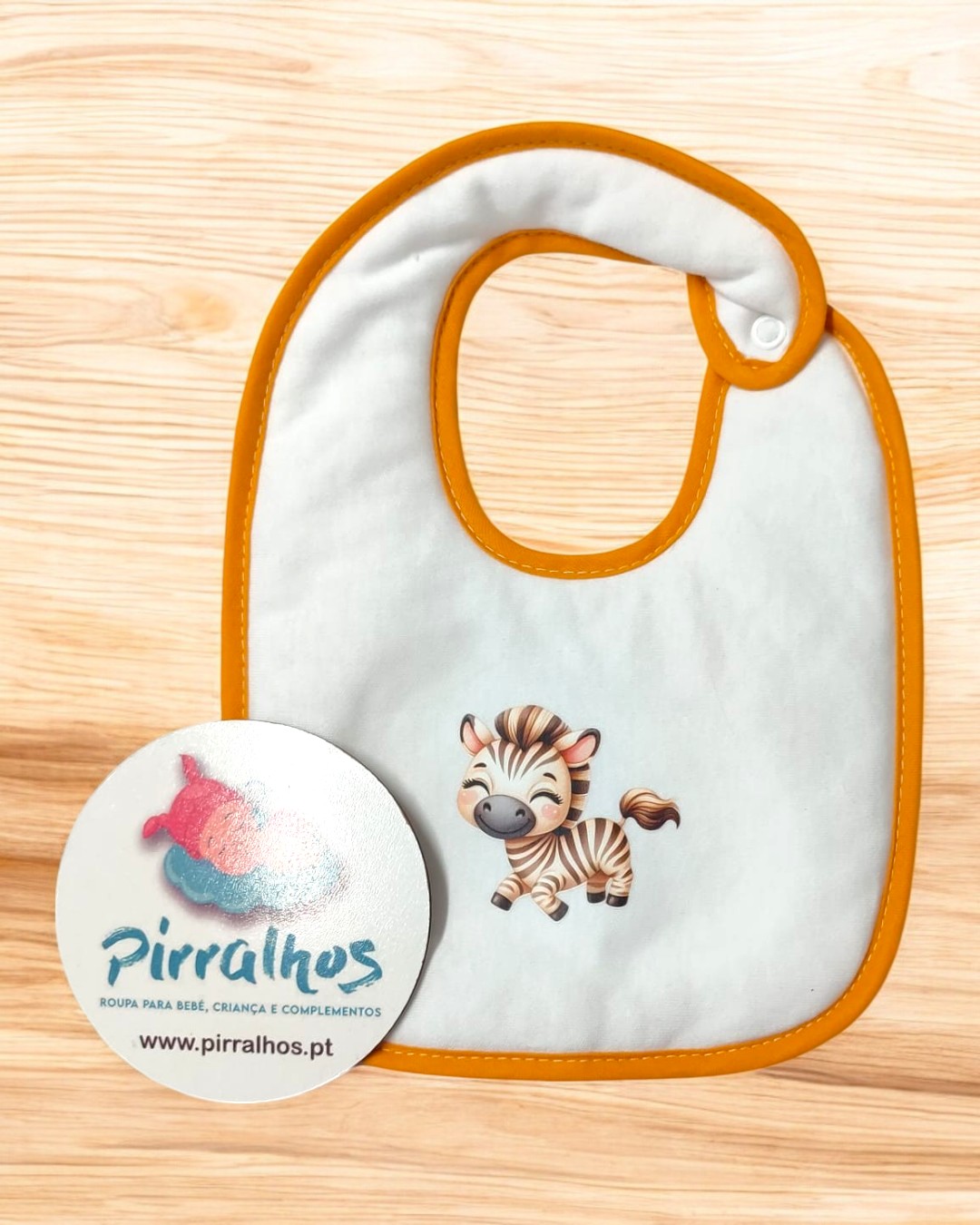 Babete branco com borda laranja e estampa de zebra, sobre superfícies de madeira clara, com cartão da marca Pirralhos.
