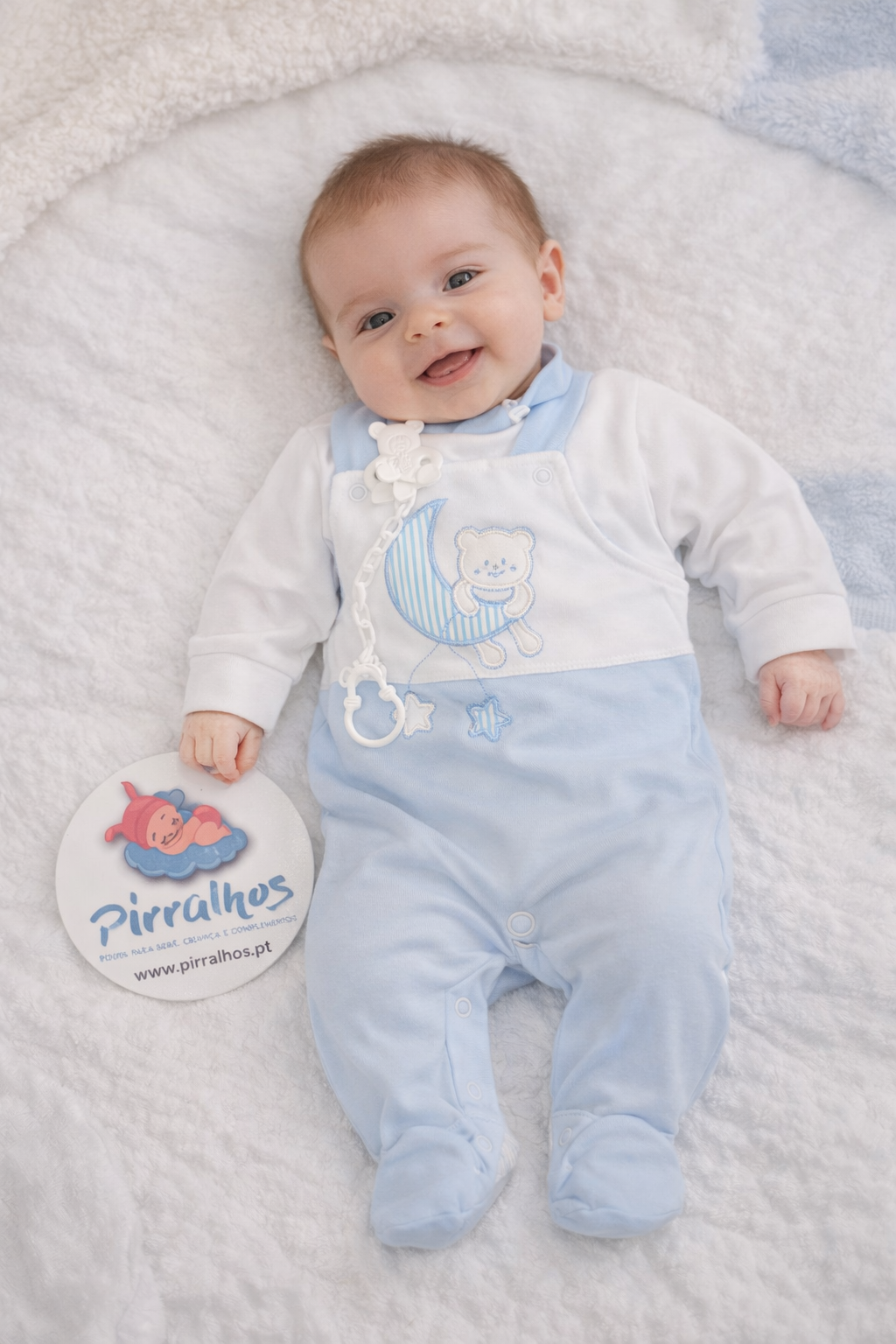 Bebé com macacão azul claro e branco deitado numa manta branca a segurar cartão com o texto Pirralhos.