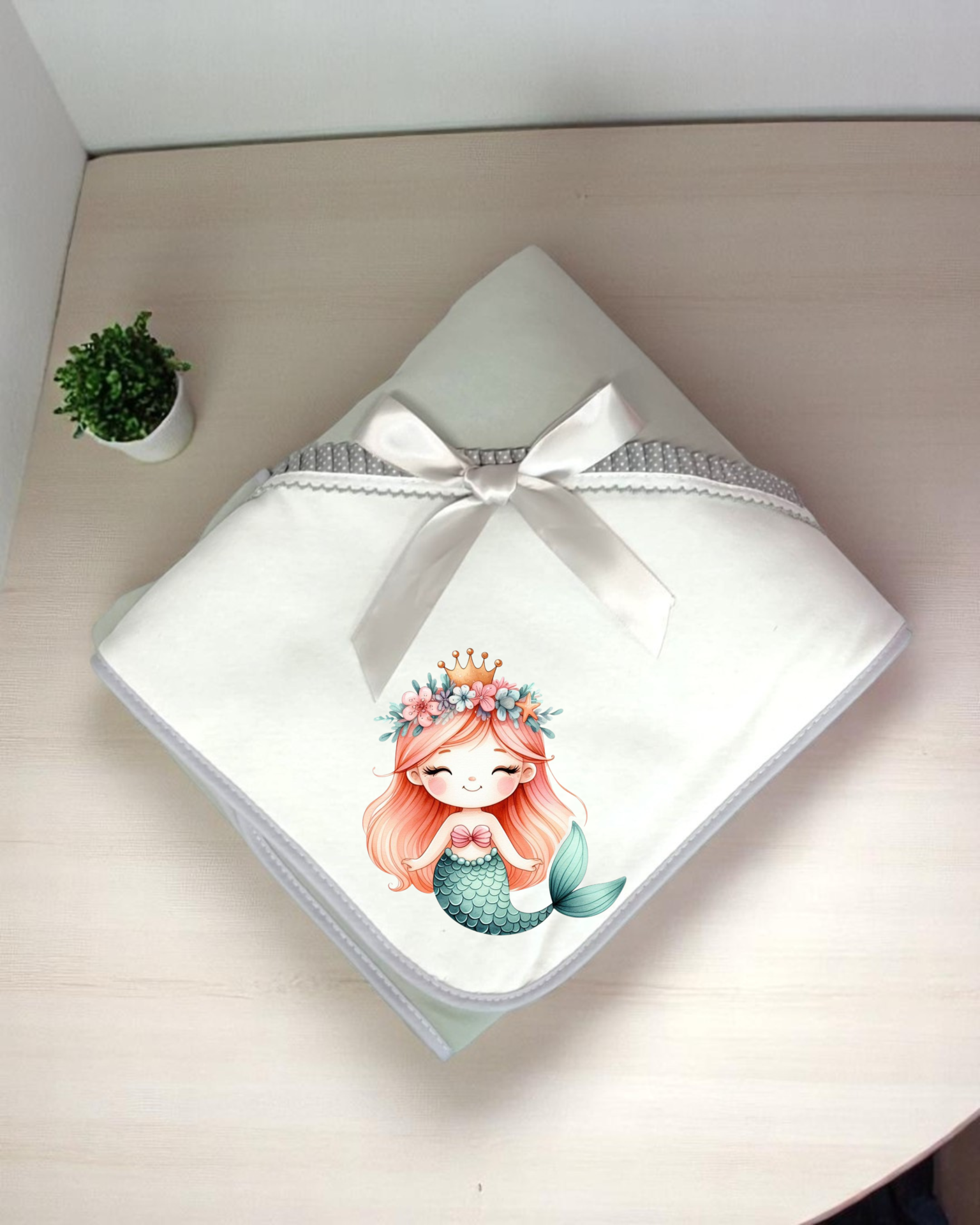 manta branca com sereia e laço de cetim sobre superfície de madeira clara ao lado de planta