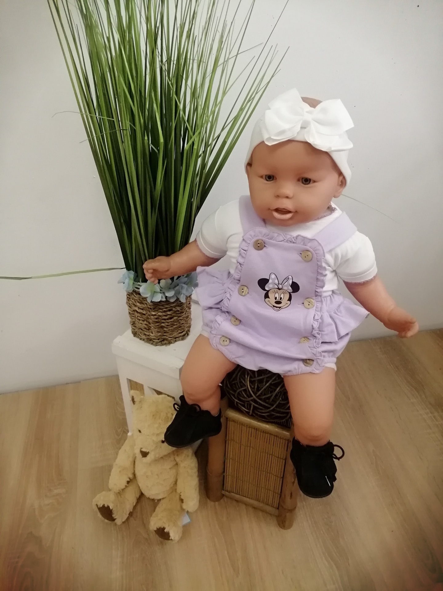 Boneca bebé com roupa lilás e branca, sentada em cadeira pequena com urso de peluche e planta ao lado