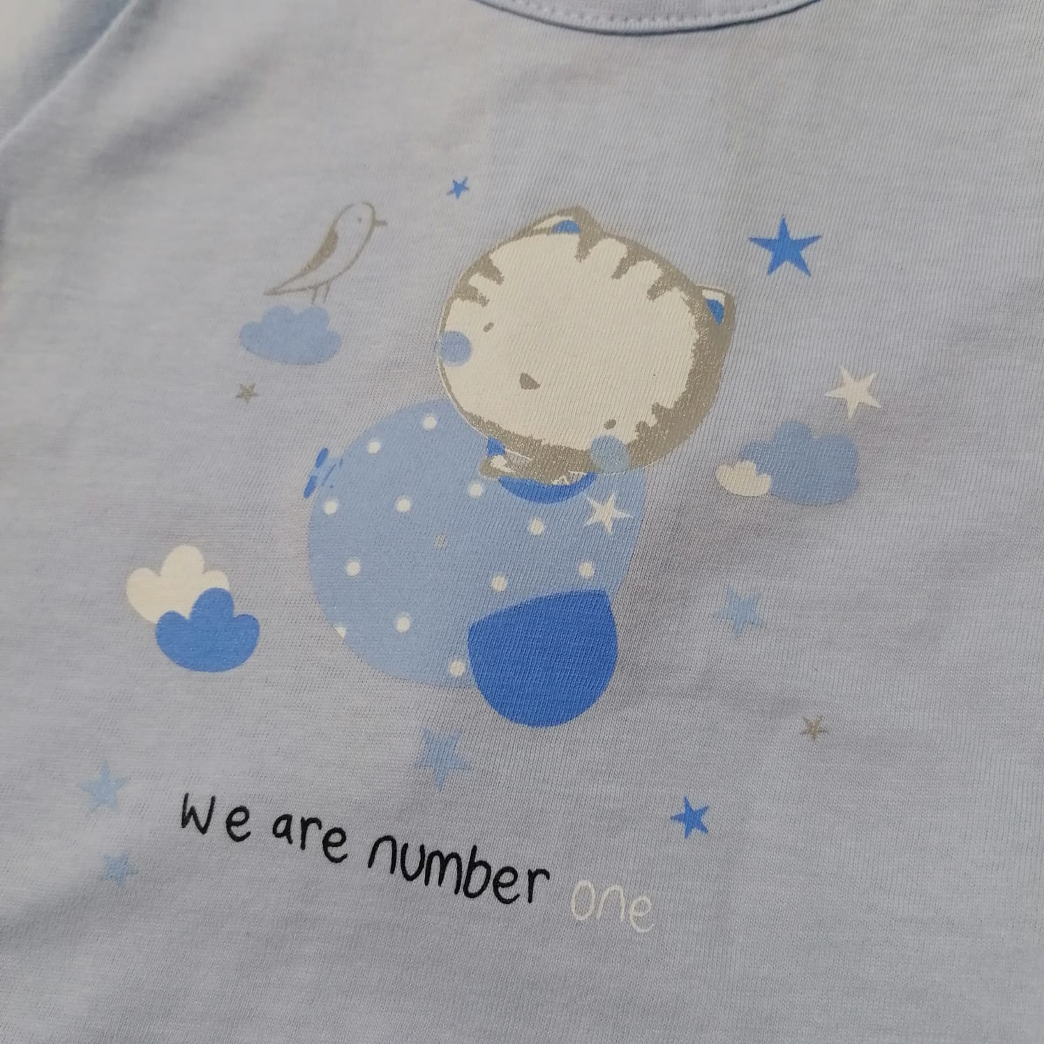 Camisola azul claro com desenho de gato, estrelas e nuvens e texto 'We are number one'.