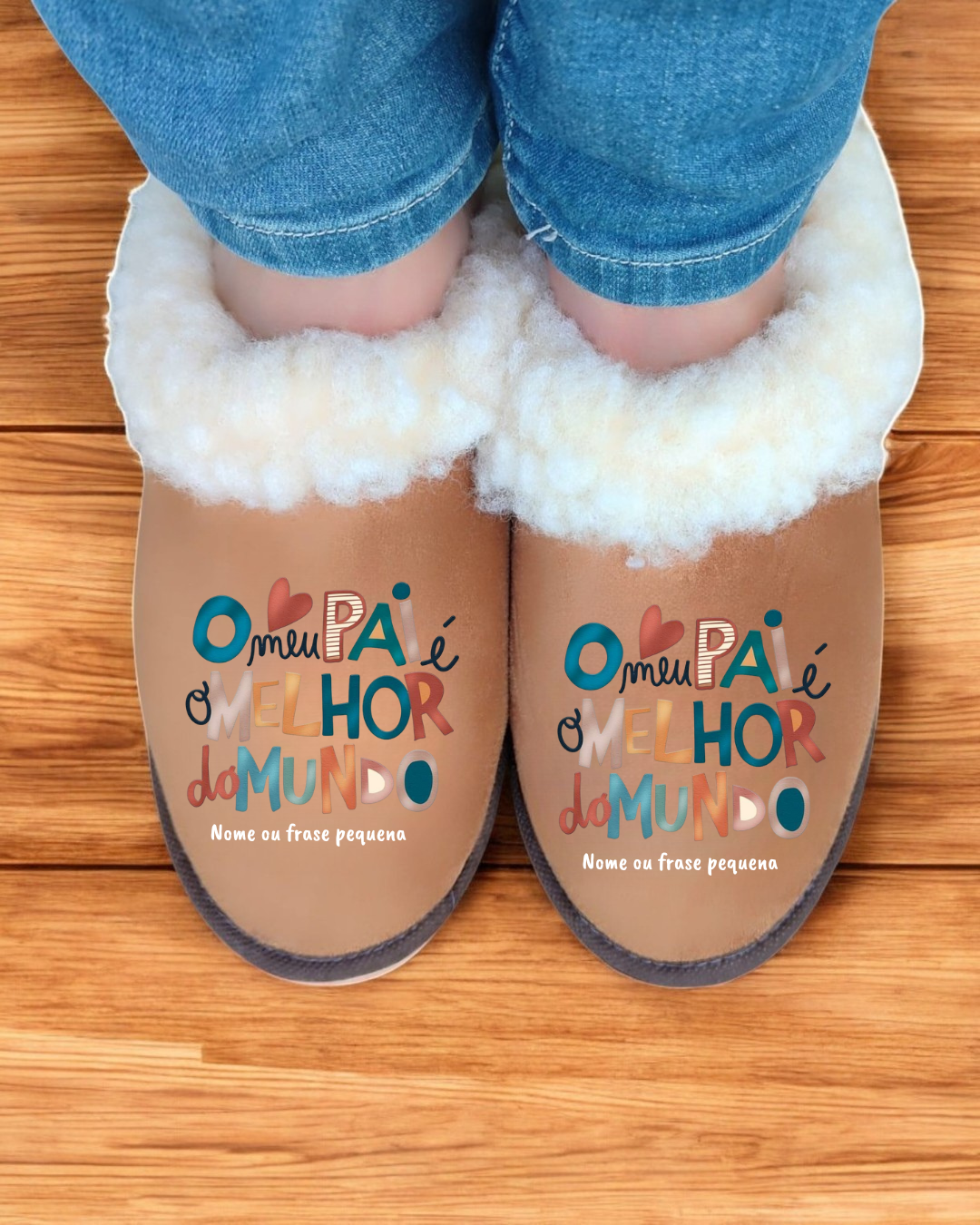 Pantufas castanhas forradas a lã branca com texto colorido no topo, usadas com jeans, em chão de madeira