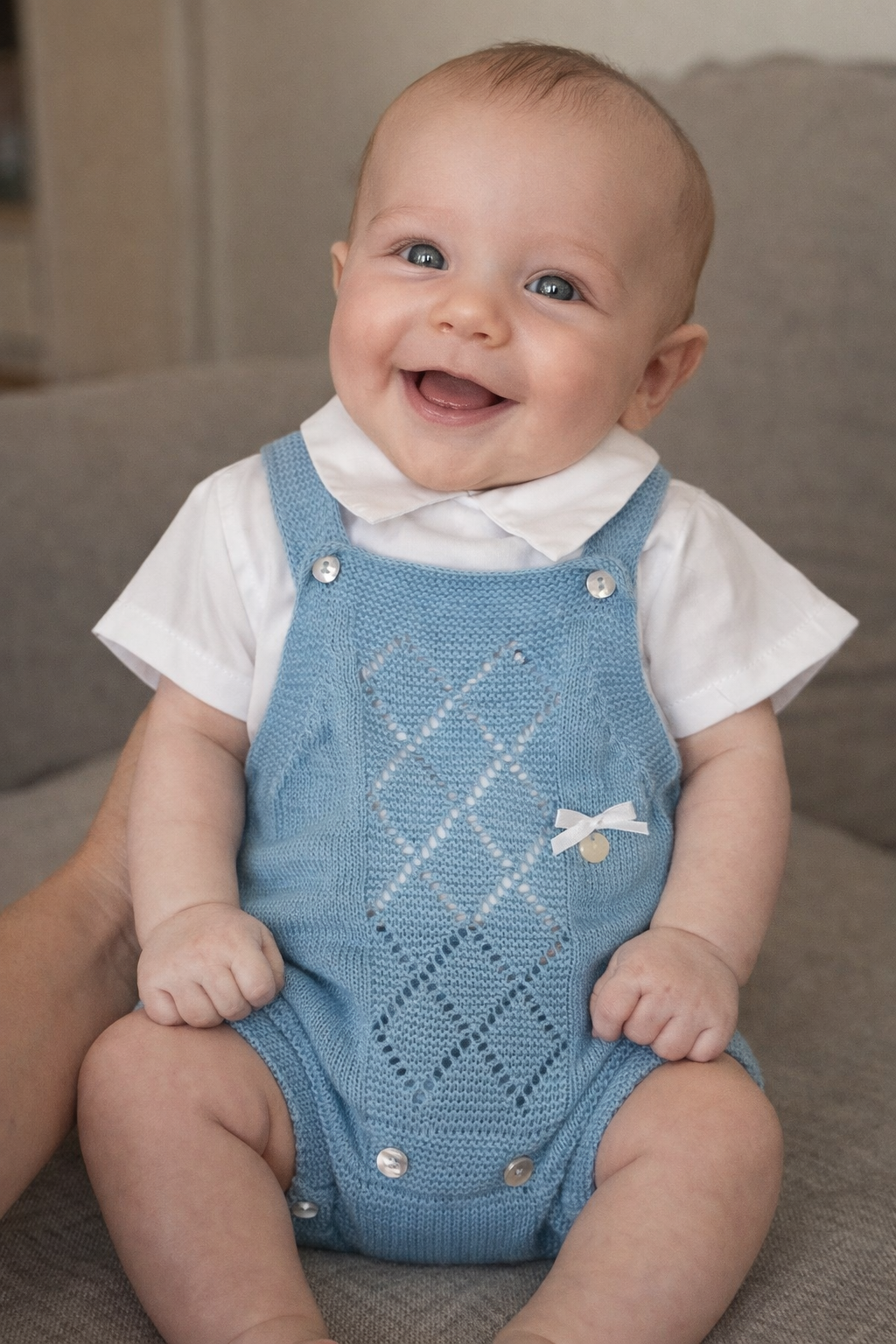 Bebé sorridente com jardineira azul de malha e body branco sentado em sofá cinzento
