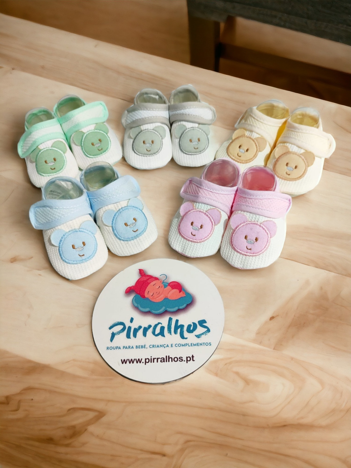 Cinco pares de sapatinhos de bebé coloridos com ursinhos e placa com logotipo pirralhos sobre mesa de madeira