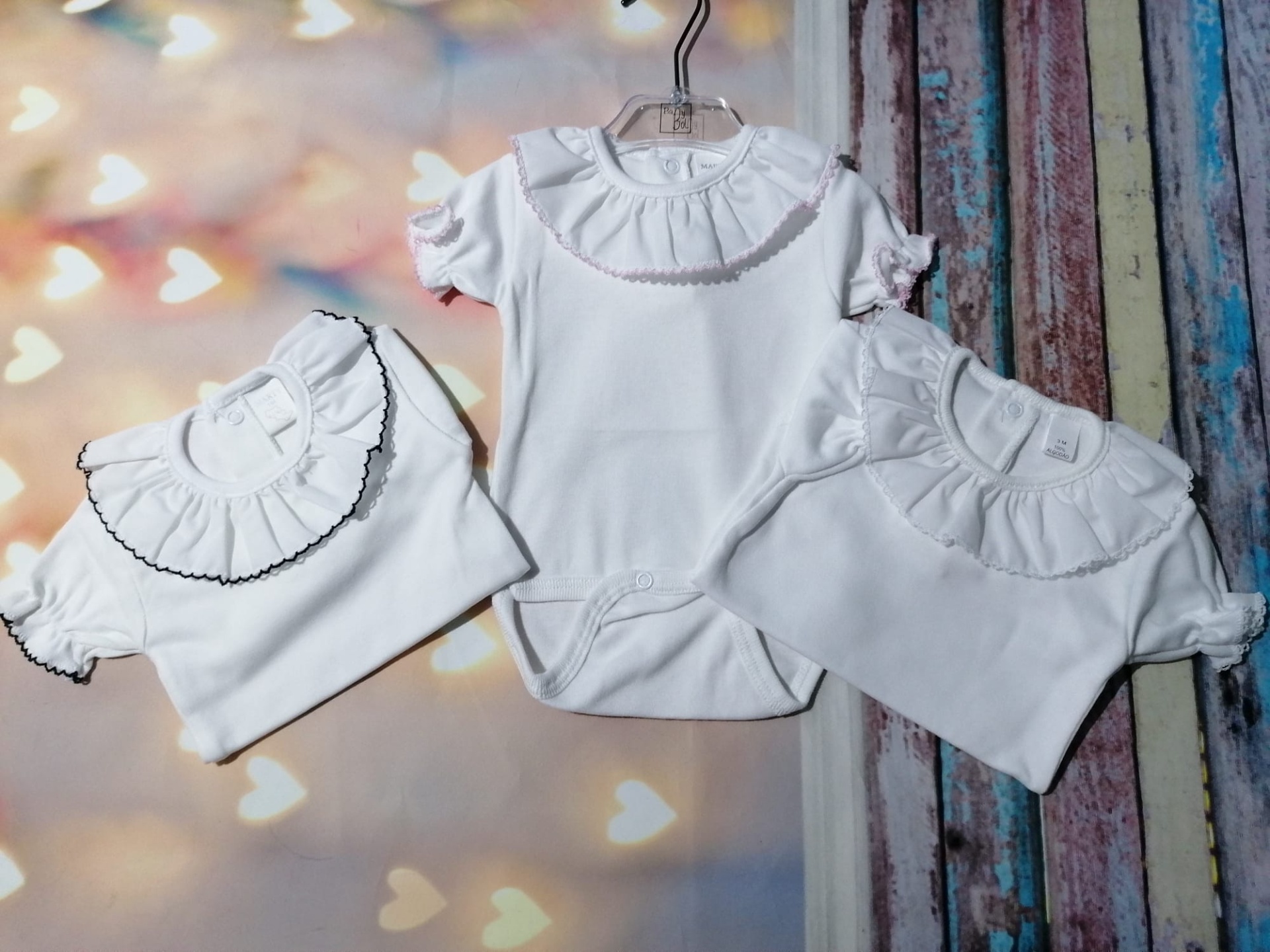 Três bodies de bebé brancos com gola rodada e detalhes franzidos, um pendurado e dois dobrados sobre superfícies com luzes desfocadas e madeira colorida.