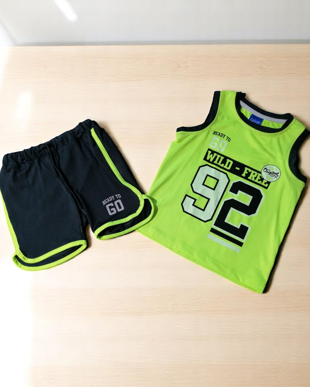 Conjunto de roupa infantil verde e preta com texto e números, sobre superfície de madeira clara