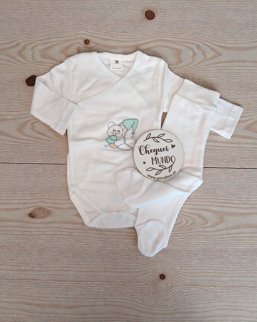 Conjunto branco para bebé com bordado de ursinho e lua, etiqueta com texto sobre madeira clara