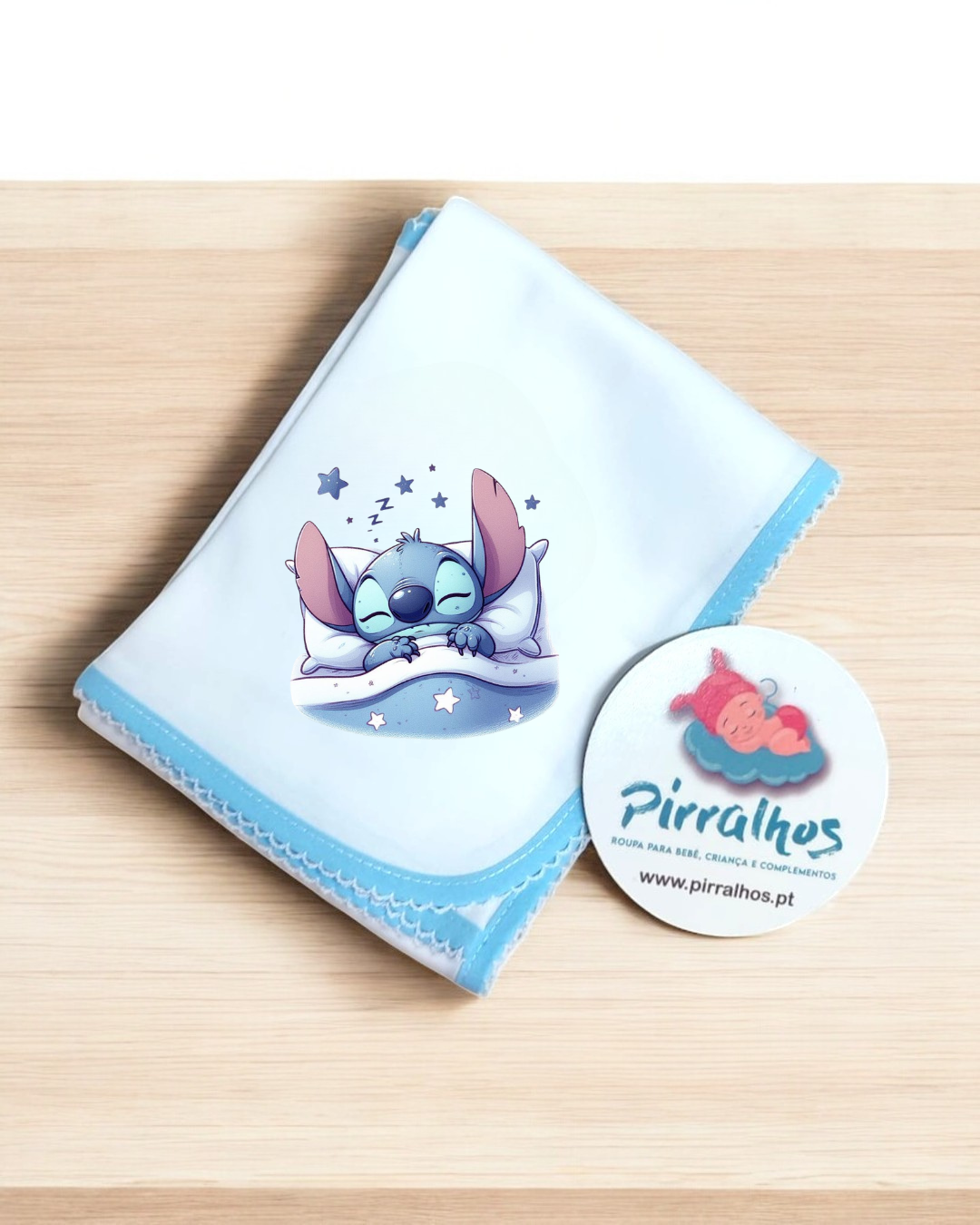 Fralda de bebé branca com borda azul e personagem Stitch a dormir, em mesa de madeira clara, com etiqueta branca redonda ao lado.