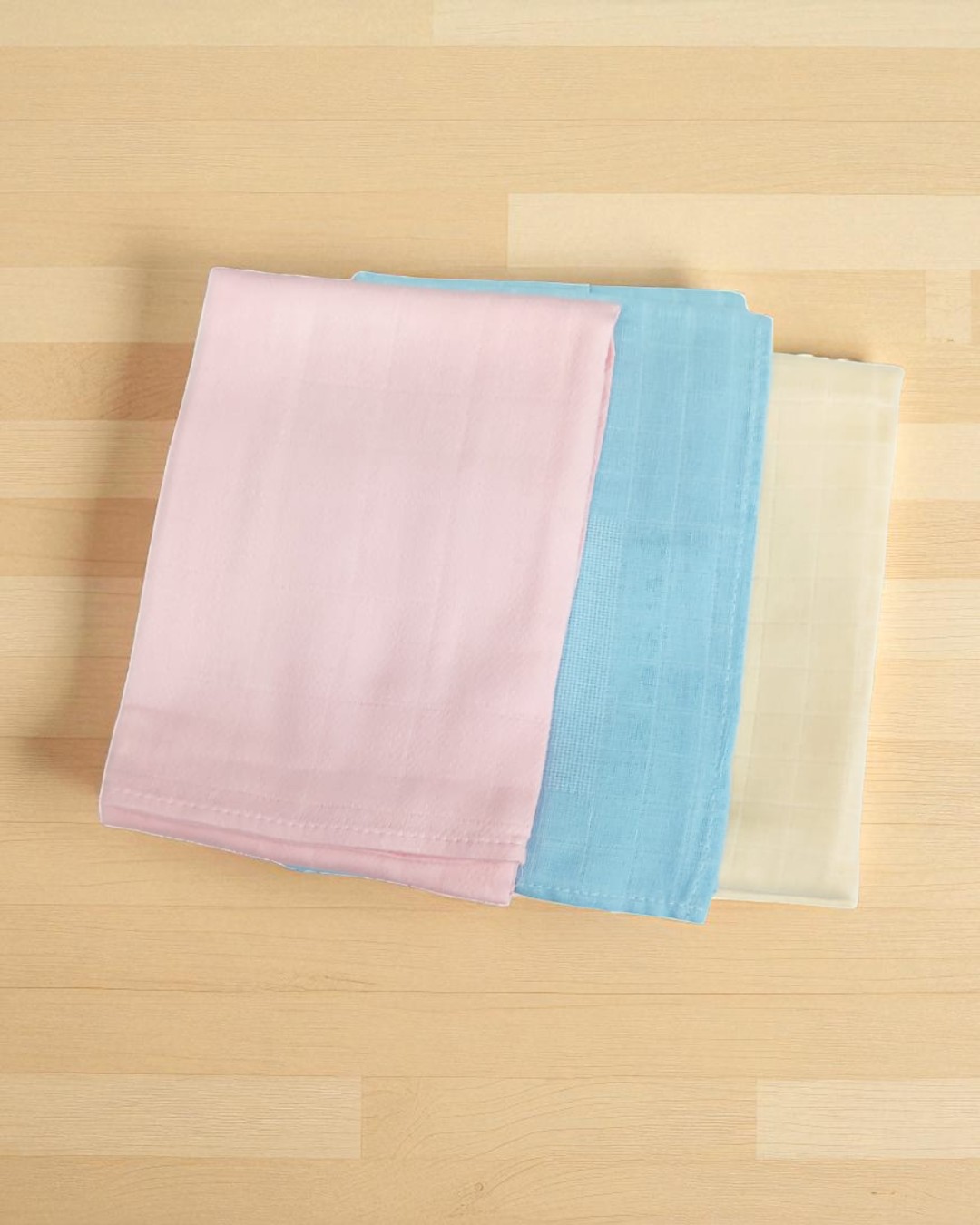 Três panos dobrados em tecido liso de cores pastel sobre madeira clara