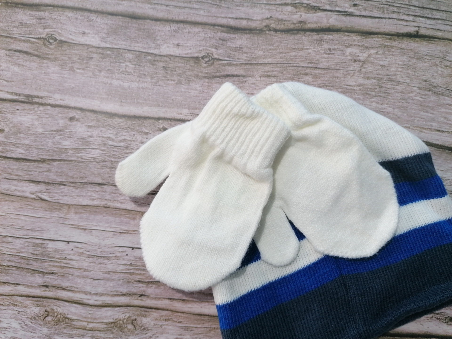 Gorro e luvas de malha branca com riscas azuis e pretas sobre superfície de madeira.