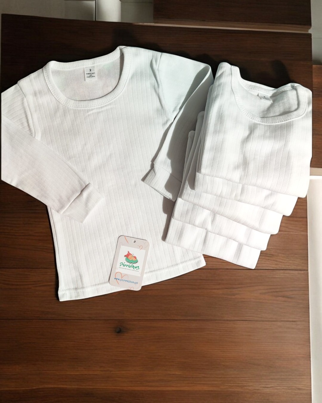 Camisolas térmicas brancas para criança em tecido canelado com etiqueta 'Pirralhos' sobre mesa de madeira