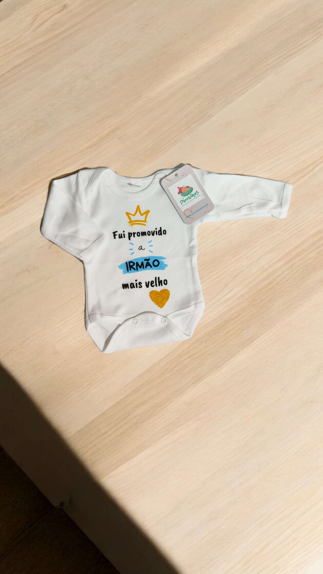 Body de bebé branco com frase em várias cores e etiqueta de marca, sobre mesa de madeira clara