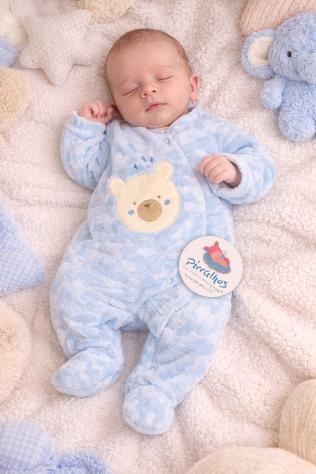 Bebé a dormir vestido com pijama azul claro com urso e etiqueta Pirralhos sobre mantas e almofadas.