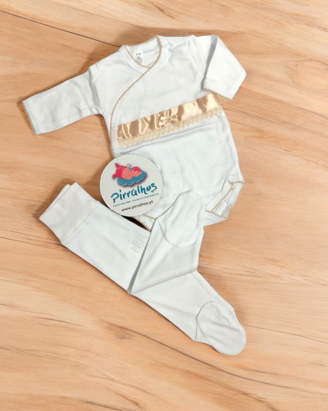 Conjunto de roupa para bebé branco com faixa dourada e calças com pés num chão de madeira clara