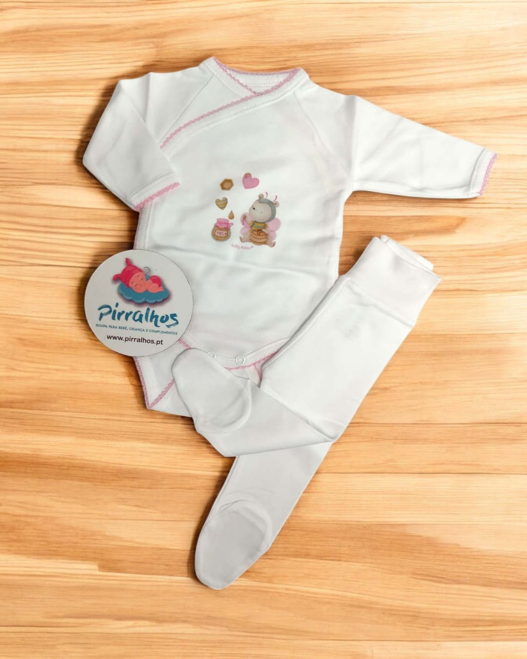 Conjunto de bebé branco com detalhes rosa e ilustração, calças com pés, etiqueta Pirralhos, fundo madeira clara