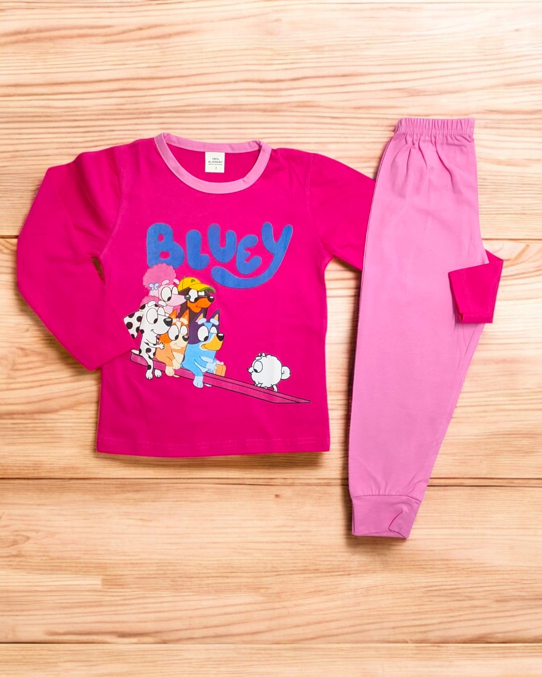 Conjunto de roupa infantil com camisola e calças rosa com estampa Bluey sobre madeira clara