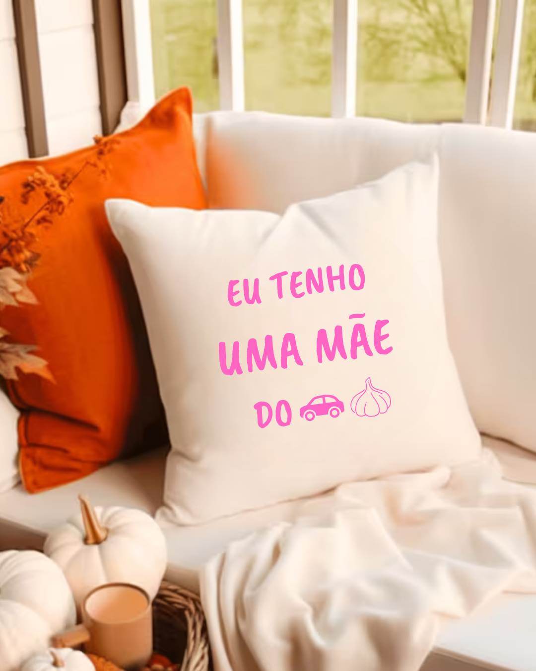 Almofada branca com texto rosa e ícones, rodeada por decoração aconchegante em tons de branco e laranja