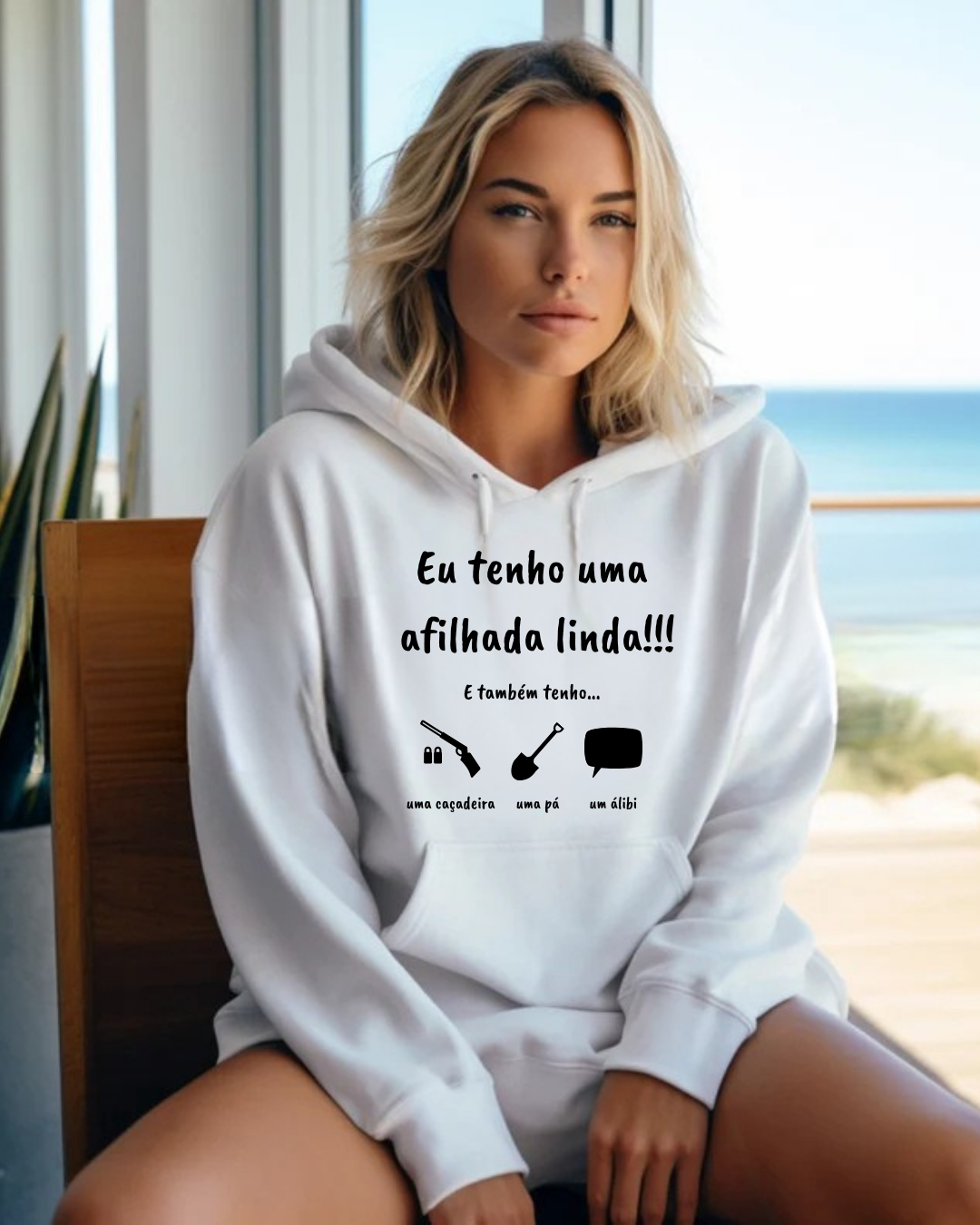 Mulher a usar casaco branco com capuz com texto em português e ícones, sentada em cadeira junto a uma janela com vista para o mar.