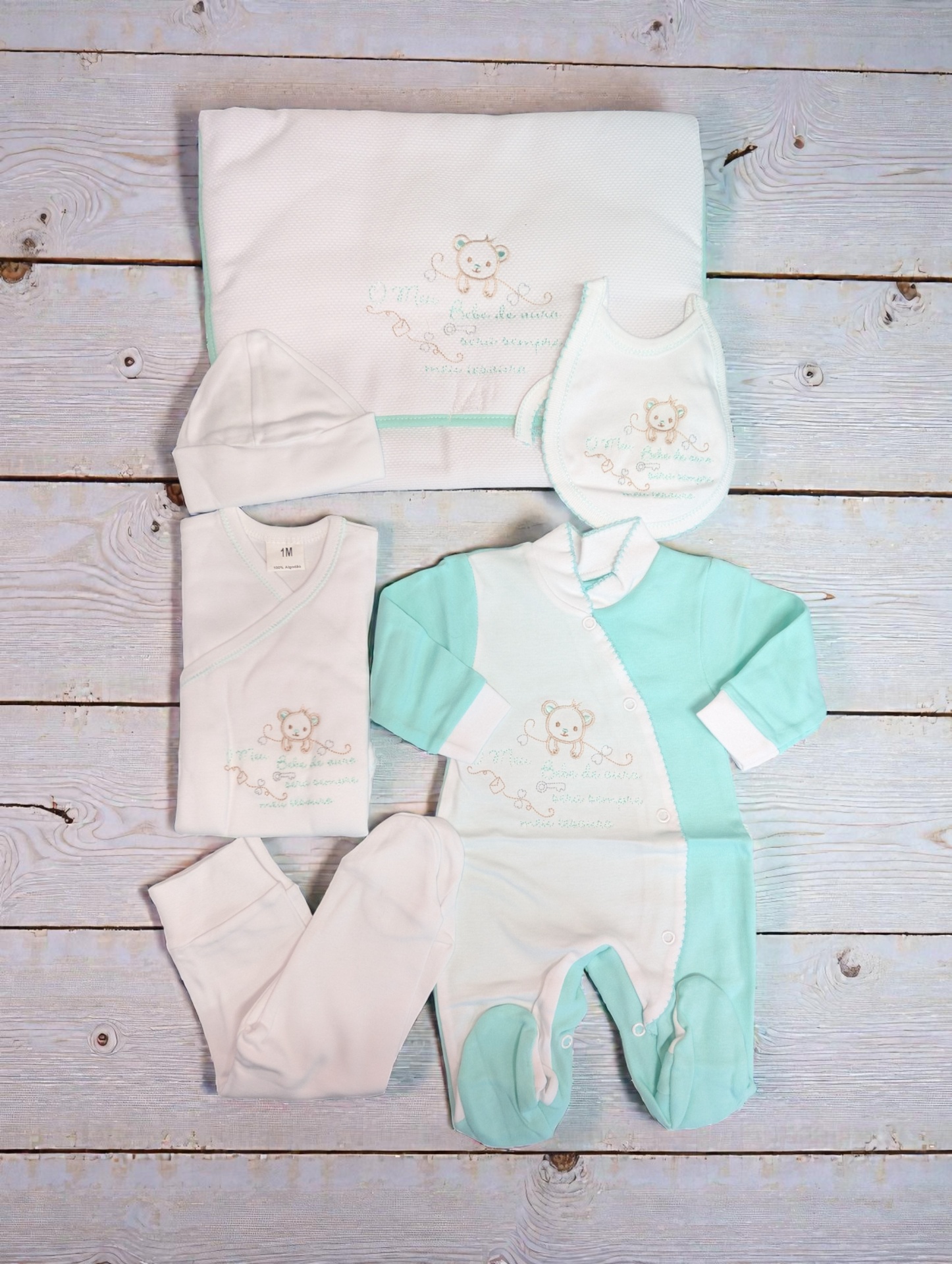Conjunto de roupa de bebê branco e verde-água com bordado de ursinho sobre madeira clara
