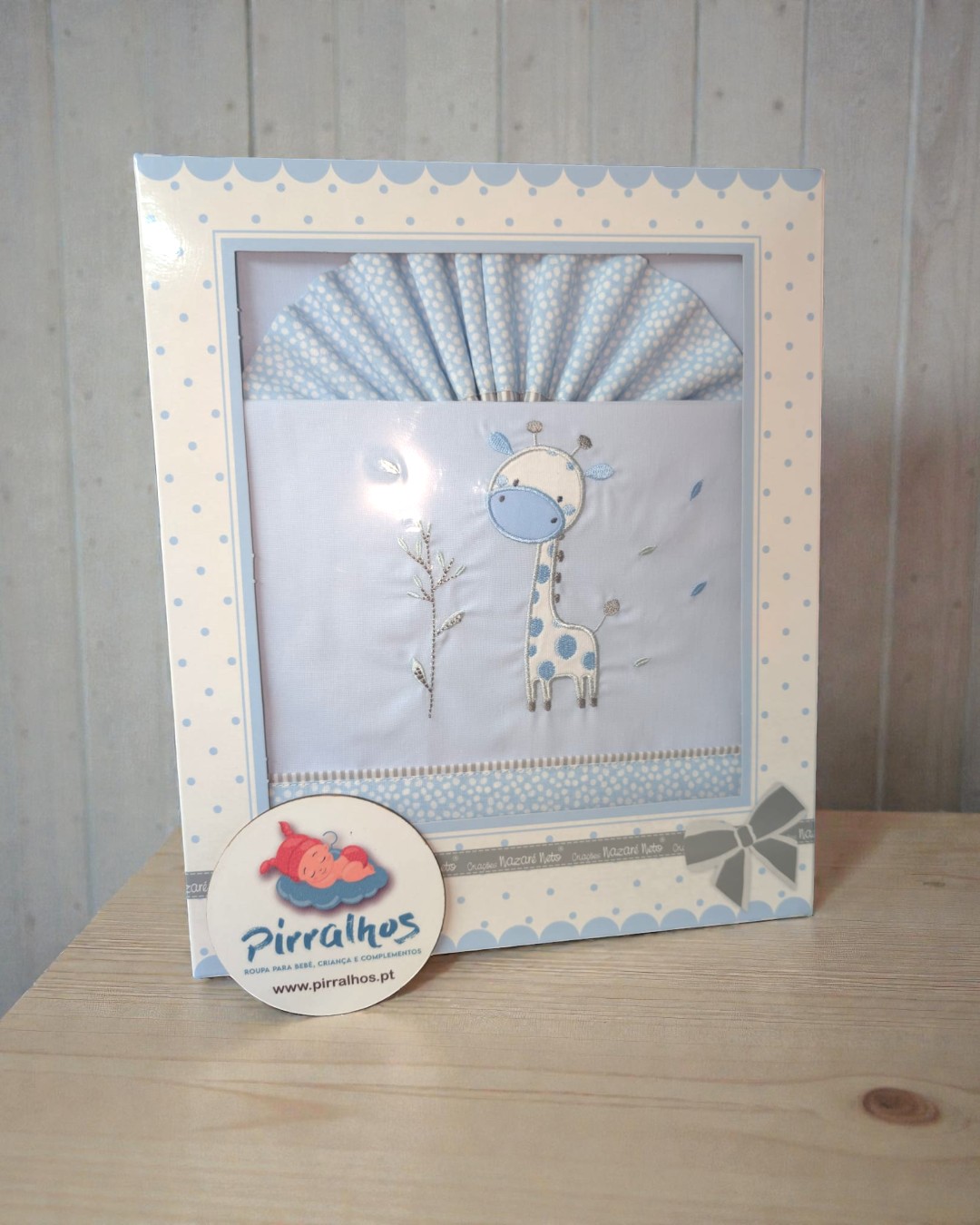 Fralda de bebé branca com bordado de girafa azul e bege numa caixa decorada com bolinhas azuis e bege.