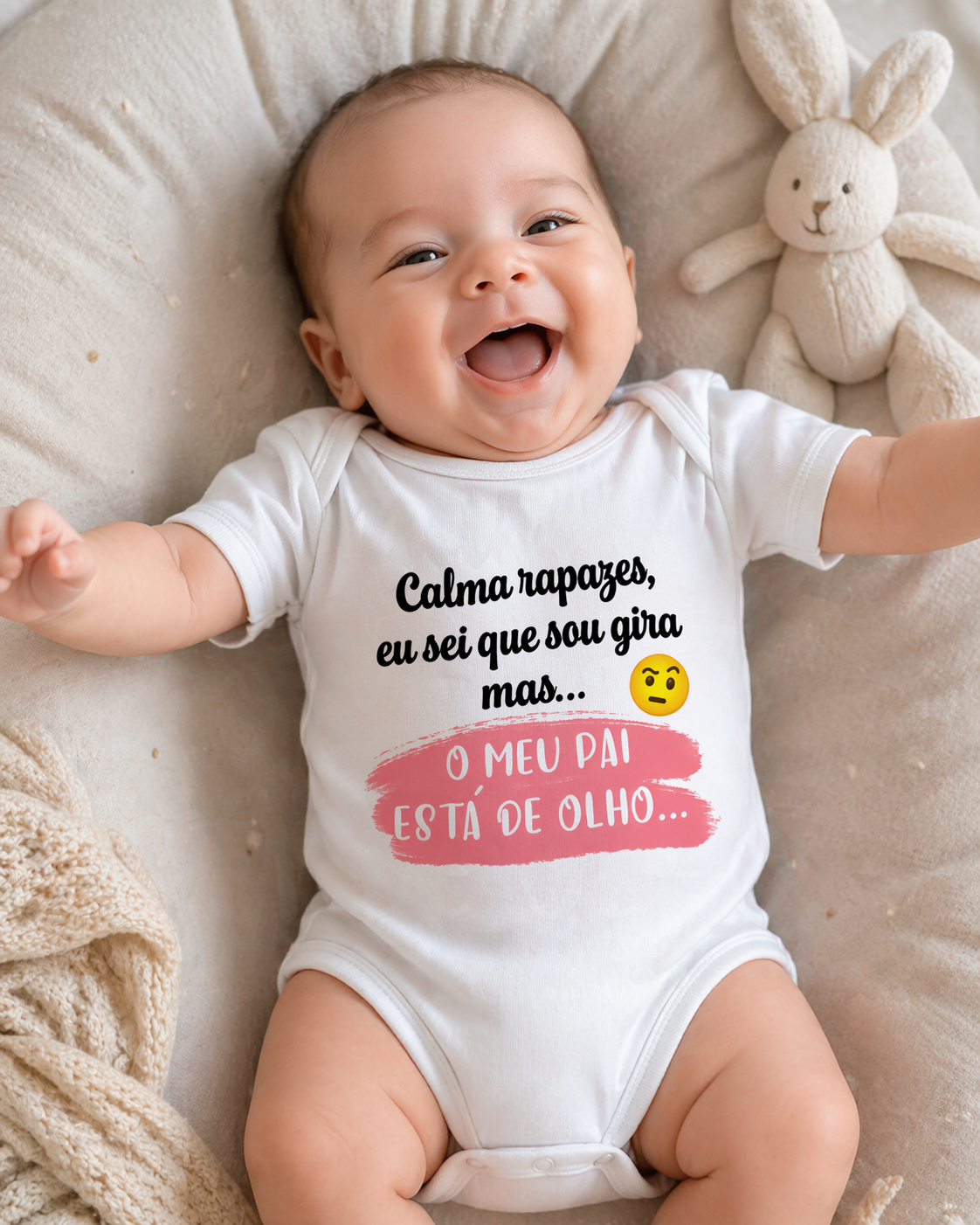 Bebé com body branco e texto estampado deitado em almofada bege ao lado de peluche coelho creme
