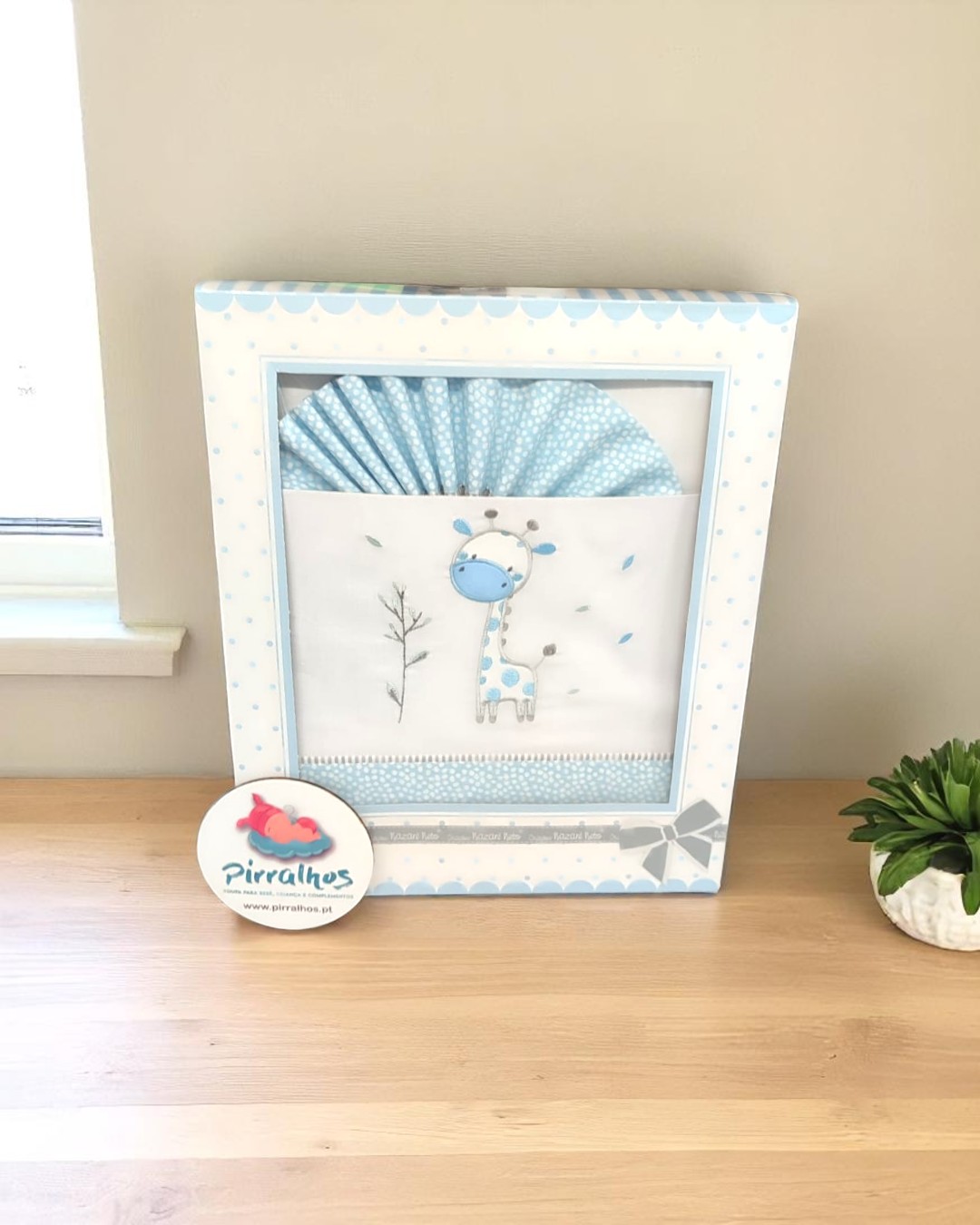 Conjunto de roupa de bebé azul com girafa bordada numa caixa decorativa com padrão de pontos azuis.