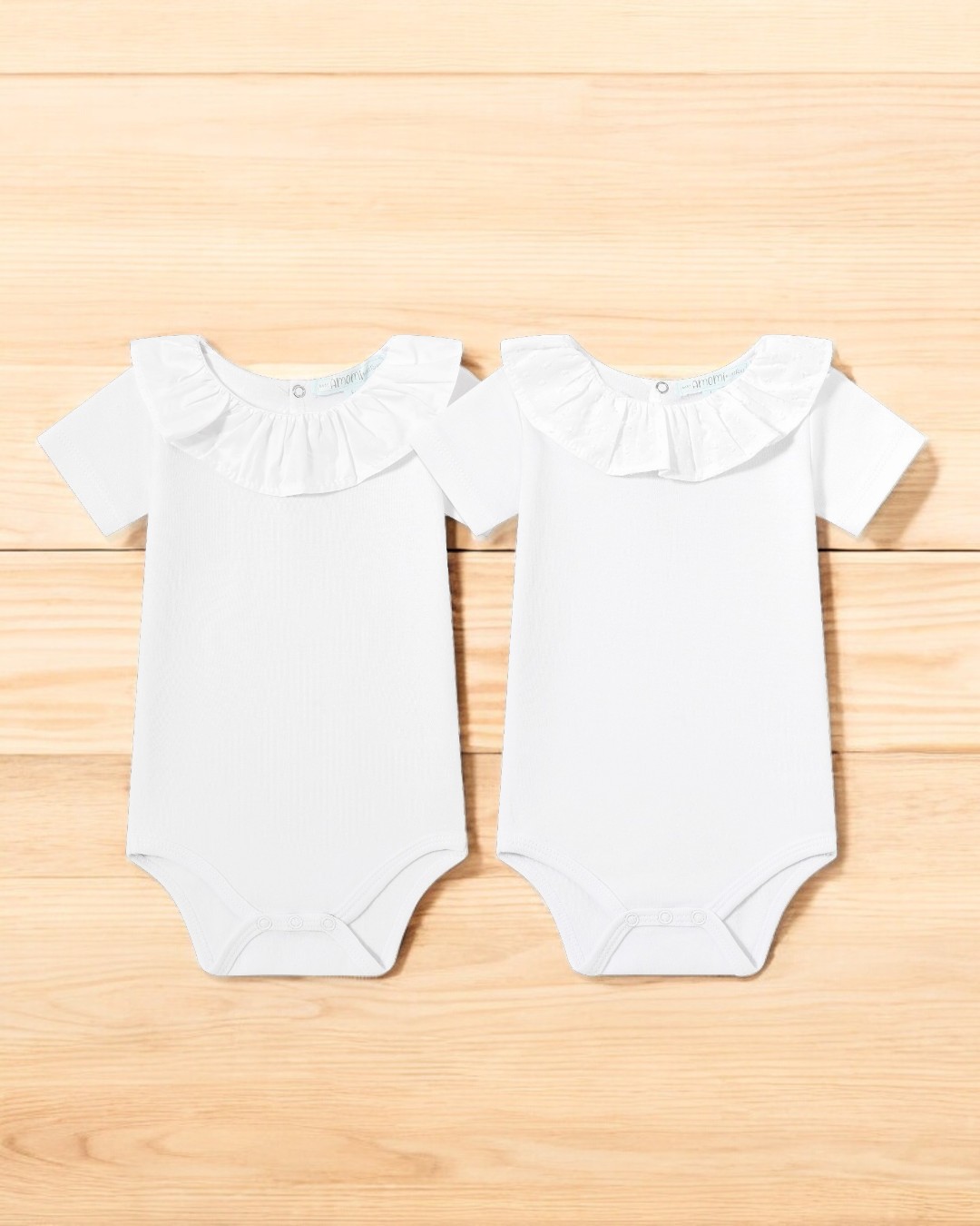 Dois bodies brancos de bebé com gola de folhos em tecido, sobre madeira clara.
