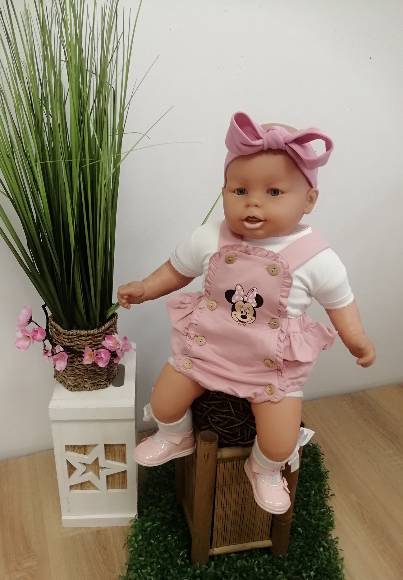 Boneca bebé com roupa rosa da Minnie e faixa de cabelo sentada em cadeira ao lado de vaso com plantas
