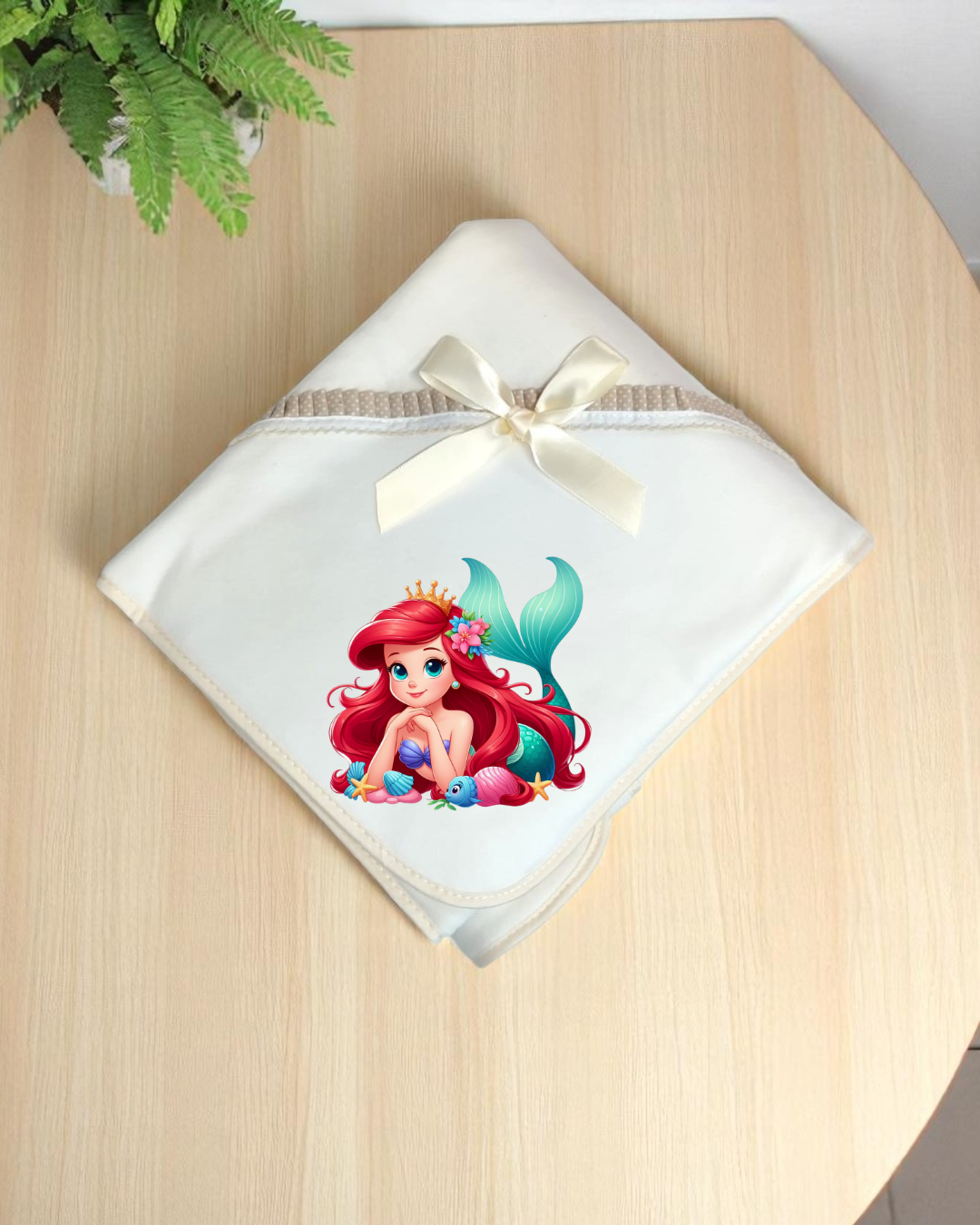 Toalha de bebé branca com laço creme e estampa de sereia colorida sobre mesa de madeira clara com planta ao lado