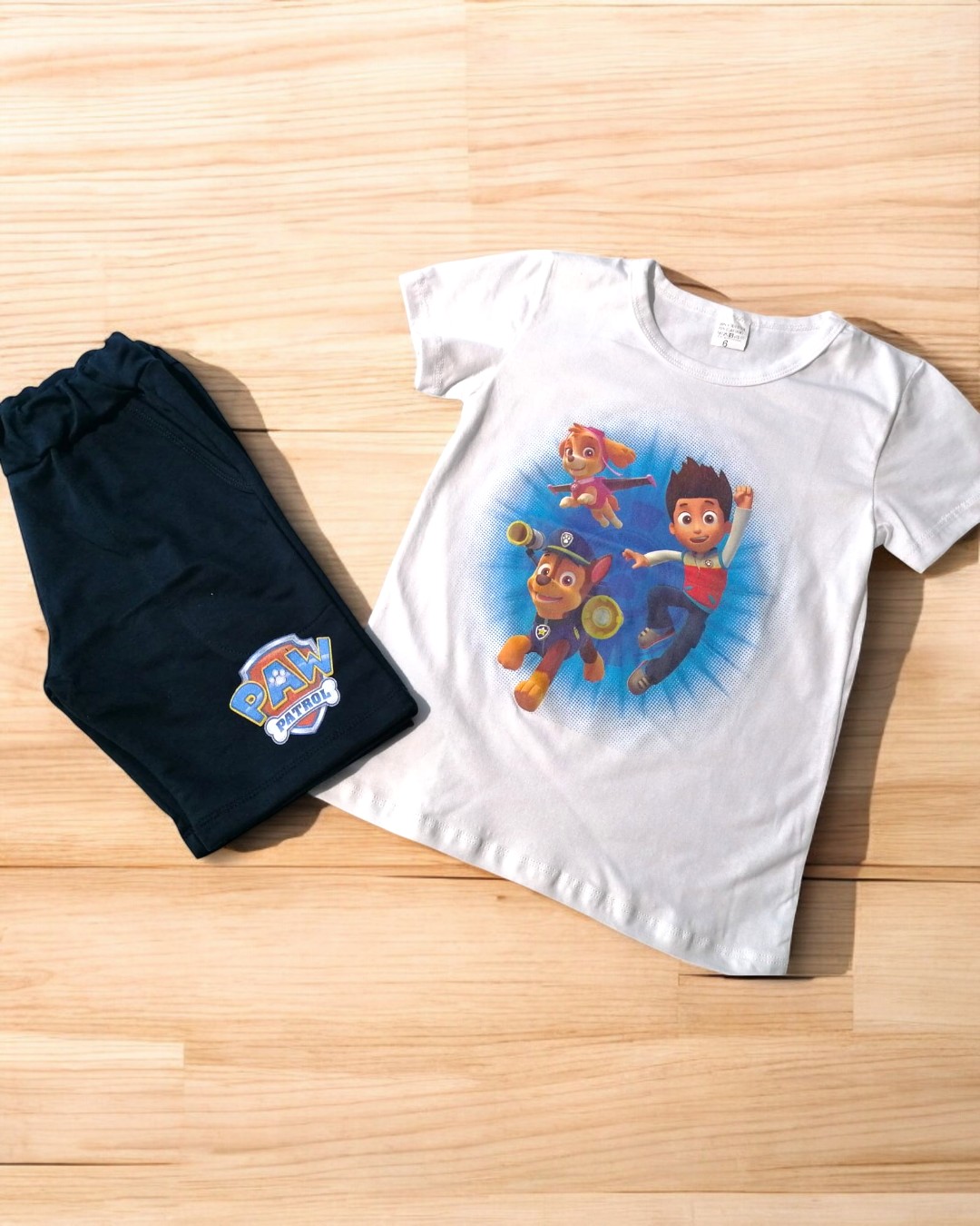 Conjunto infantil de t-shirt branca com personagens da Patrulha Pata e calças pretas, dispostos sobre chão de madeira clara.